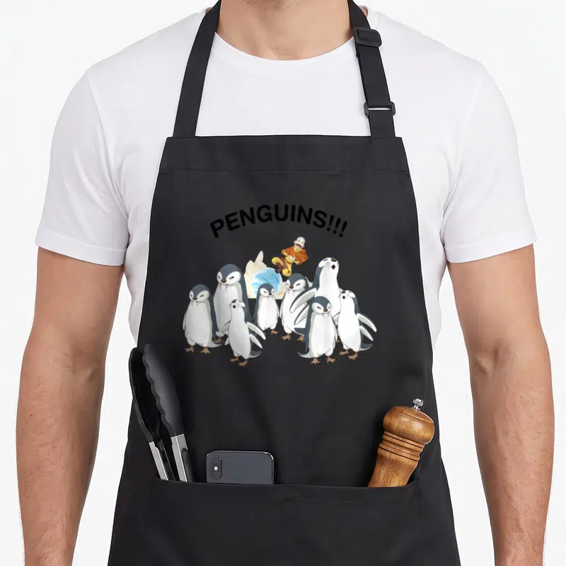 Aang penguin aprons - Arched PENGUINS!!! text over a group of penguins and a falling character in orange robes — shown on a black cotton canvas apron against a white background
