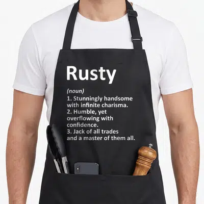 Rusty father day aprons - White text defining Rusty as handsome, humble, and a jack of all trades stacked vertically — shown on a black cotton canvas apron against a white background