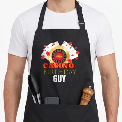 Casino chef outfit with apron - Roulette wheel, fanned cards, chips, and stacked Casino Birthday Guy text in red gold — shown on a black cotton canvas apron against a white background