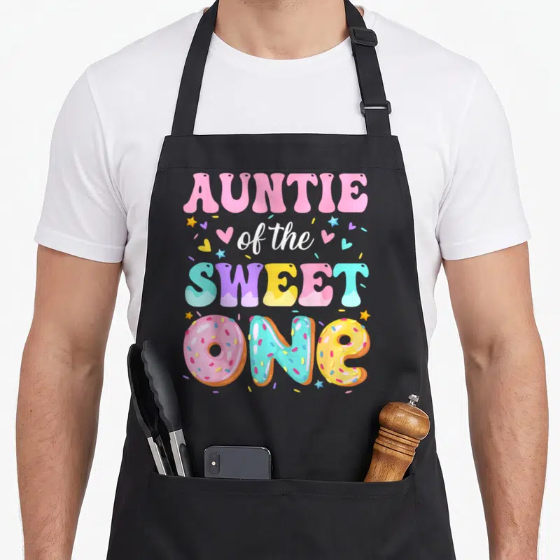 Auntie donut apron - Stacked text Auntie of the Sweet One with donut shaped letters and scattered sprinkles — shown on a black cotton canvas apron against a white background