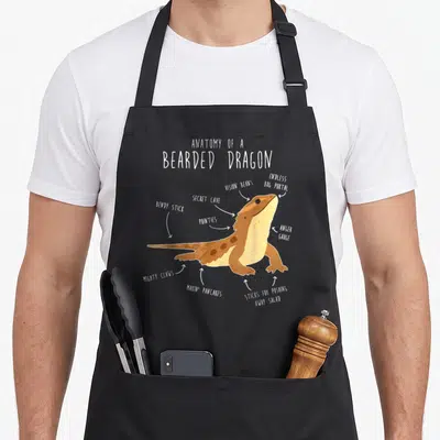 Dragon anatomy apron - Cartoon bearded dragon diagram with labels vision beans and makin pancakes — shown on a black cotton canvas apron against a white background