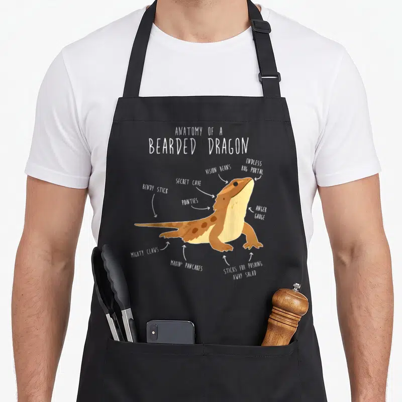 Dragon anatomy apron - Cartoon bearded dragon diagram with labels vision beans and makin pancakes — shown on a black cotton canvas apron against a white background