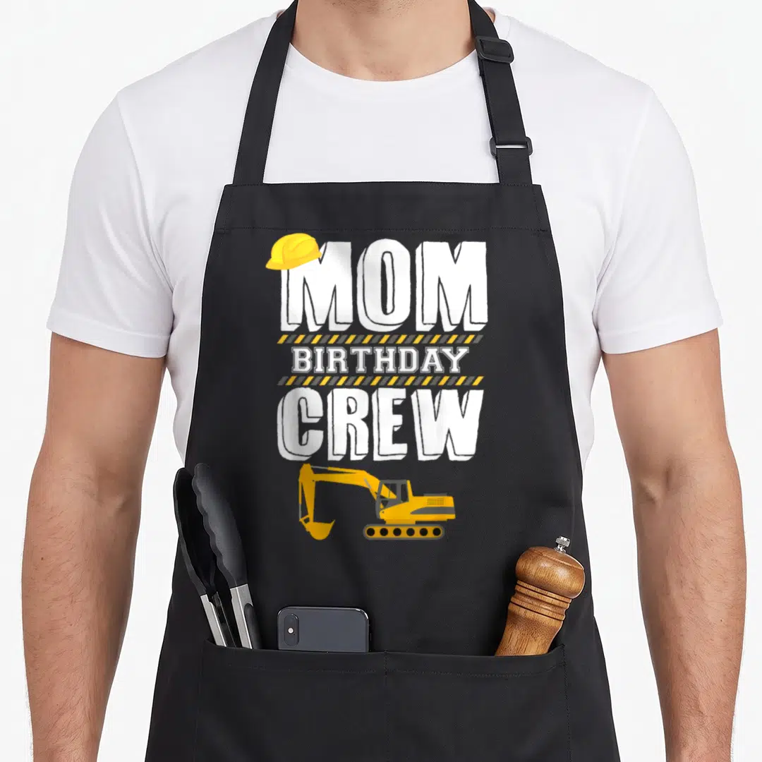 Hosting construction worker apron - MOM BIRTHDAY CREW text with hard hat, hazard stripes, and excavator illustration — shown on a black cotton canvas apron against a white background