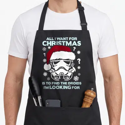 Storm christmas apron - Cartoon stormtrooper in red santa hat with light blue holiday pun text and scattered snowflakes — shown on a black cotton canvas apron against a white background