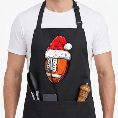 Christmas sports team aprons - Cartoon brown football with white laces wearing a red and white Santa hat — shown on a black cotton canvas apron against a white background