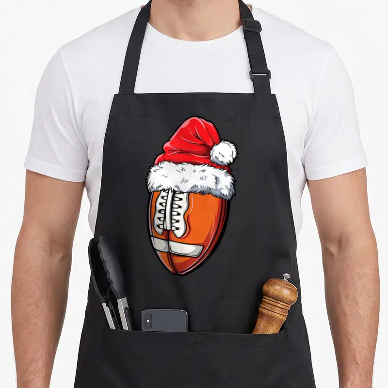 Christmas sports team aprons - Cartoon brown football with white laces wearing a red and white Santa hat — shown on a black cotton canvas apron against a white background