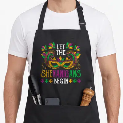Celebration mardi gras aprons - Retro masquerade mask with LET THE SHENANIGANS BEGIN text and fleur-de-lis accents — shown on a black cotton canvas apron against a white background