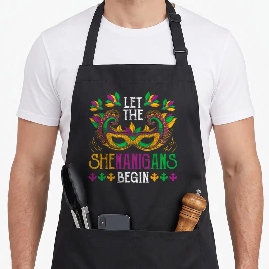 Celebration mardi gras aprons - Retro masquerade mask with LET THE SHENANIGANS BEGIN text and fleur-de-lis accents — shown on a black cotton canvas apron against a white background