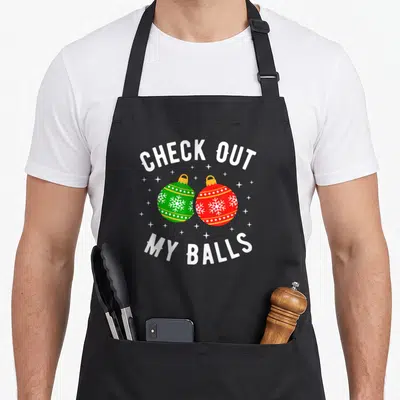 Check the dirty apron - Two cartoon Christmas ornaments in red and green with white snowflakes framed by arched white — shown on a black cotton canvas apron against a white background