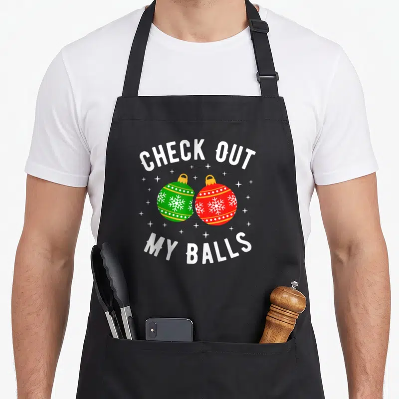 Check the dirty apron - Two cartoon Christmas ornaments in red and green with white snowflakes framed by arched white — shown on a black cotton canvas apron against a white background