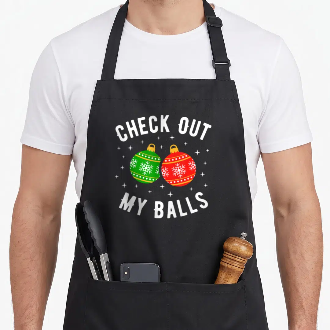Check the dirty apron - Two cartoon Christmas ornaments in red and green with white snowflakes framed by arched white — shown on a black cotton canvas apron against a white background