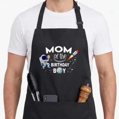 Space family aprons - Cartoon astronaut holding cake next to MOM Of the BIRTHDAY BOY text with moon — shown on a black cotton canvas apron against a white background