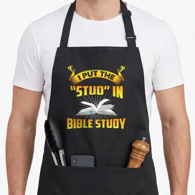 Stud bible study apron - I Put The Stud In Bible Study text above glowing open book in yellow and grey cartoon style — shown on a black cotton canvas apron against a white background