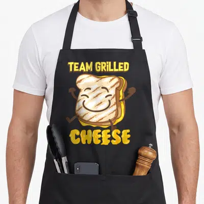 Cheese grilled lover apron - Cartoon grilled cheese sandwich running with TEAM GRILLED CHEESE text in yellow and brown — shown on a black cotton canvas apron against a white background