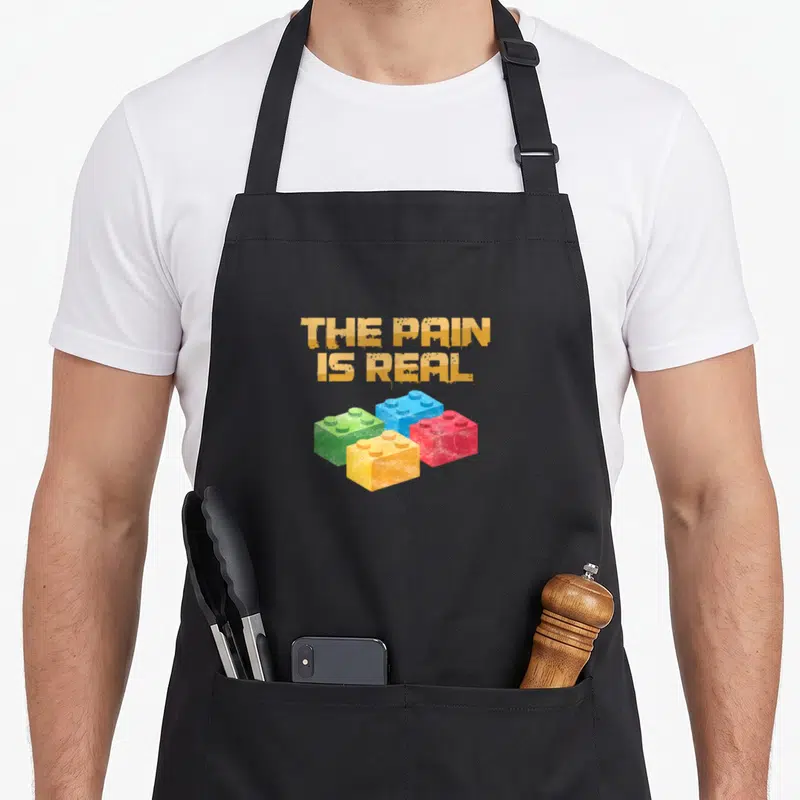 Pain block & apron - THE PAIN IS REAL text above green blue red and yellow plastic building blocks — shown on a black cotton canvas apron against a white background