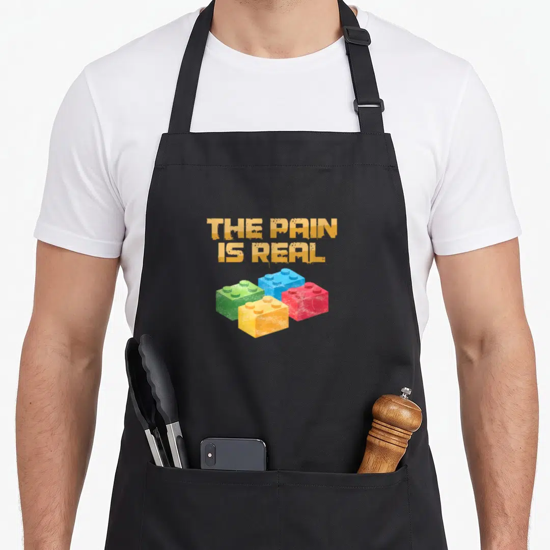 Pain block & apron - THE PAIN IS REAL text above green blue red and yellow plastic building blocks — shown on a black cotton canvas apron against a white background