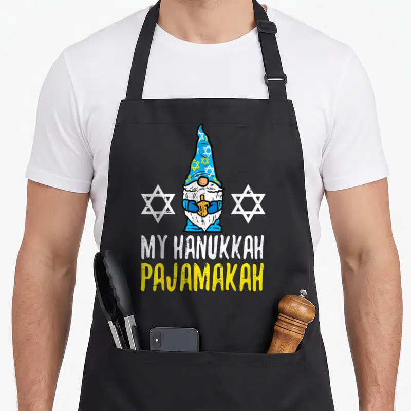 Chanukah hanukkah apron - Cartoon gnome holding dreidel between two stars with MY HANUKKAH PAJAMAKAH text — shown on a black cotton canvas apron against a white background