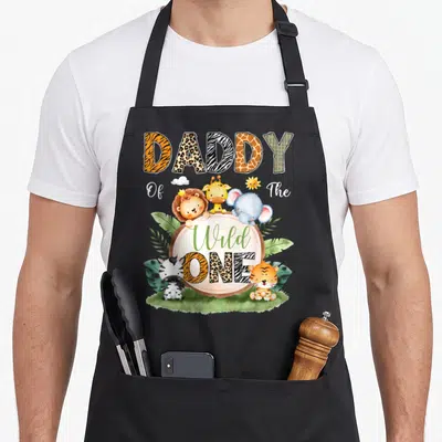 Safari daddy apron - DADDY Of The Wild ONE text with cartoon lion, giraffe, elephant, zebra, and tiger in animal print — shown on a black cotton canvas apron against a white background