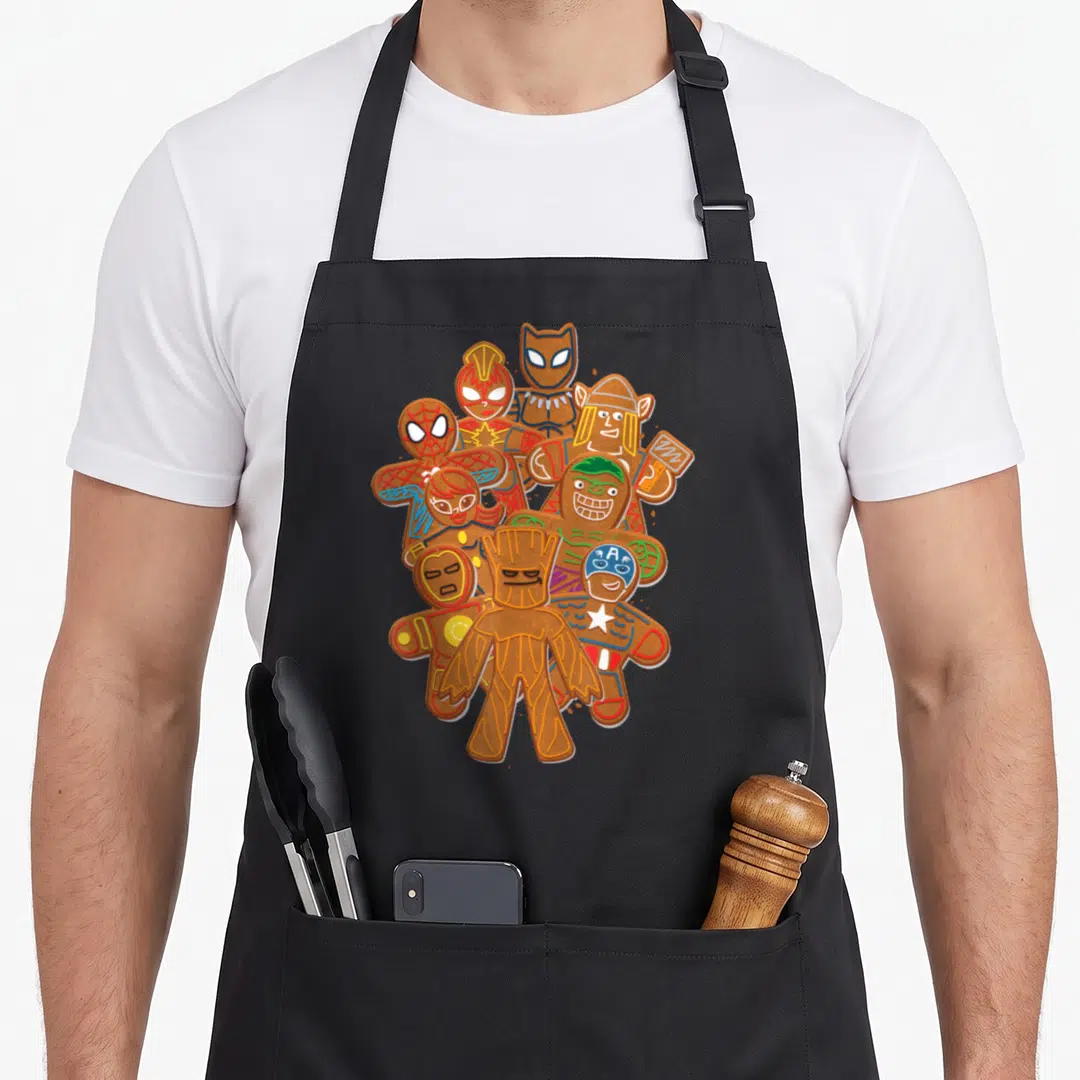 Gingerbread women's christmas aprons - Nine gingerbread superheroes including Groot in brown, red, blue, yellow — shown on a black cotton canvas apron against a white background