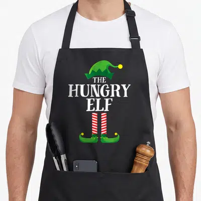 Hungry elf apron - Stacked text THE HUNGRY ELF with green hat, red white striped legs, and green shoes — shown on a black cotton canvas apron against a white background