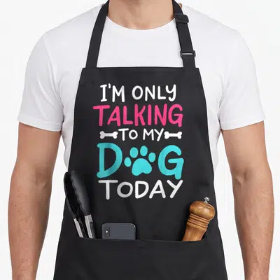 Today owner dog apron - Stacked text I'M ONLY TALKING TO MY DOG TODAY with bones and paw print in white pink and blue — shown on a black cotton canvas apron against a white background
