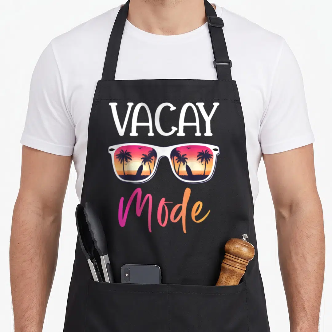 Mode sunglass summer aprons - VACAY MODE text above sunglasses reflecting a pink and orange sunset beach scene — shown on a black cotton canvas apron against a white background
