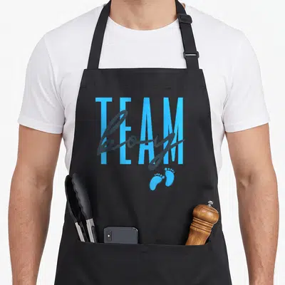 Reveal baby apron or apron - Team boy text in blue block and grey script with two footprints on black fabric — shown on a black cotton canvas apron against a white background