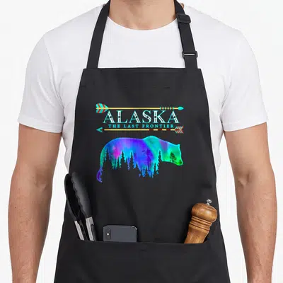 Northern bear apron - Alaska The Last Frontier text with arrows and bear silhouette filled with cyan purple aurora — shown on a black cotton canvas apron against a white background
