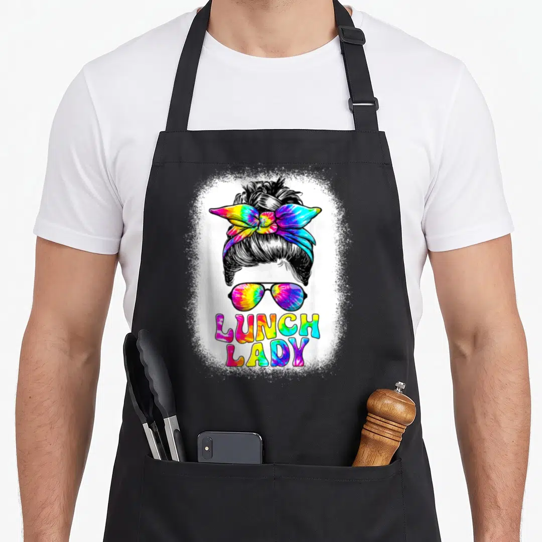 Messy lunch lady apron - Cartoon woman's head with tie-dye bow and sunglasses above LUNCH LADY text on splatter — shown on a black cotton canvas apron against a white background
