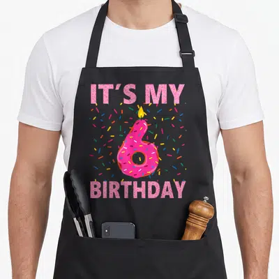 6th donut apron - Pink number six donut with candle and IT'S MY BIRTHDAY text surrounded by sprinkles — shown on a black cotton canvas apron against a white background