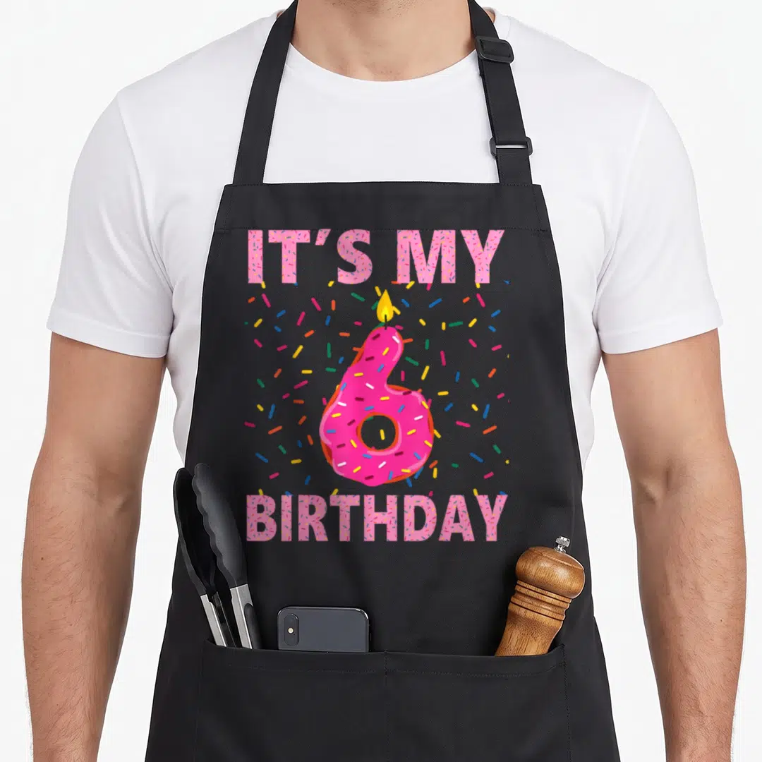6th donut apron - Pink number six donut with candle and IT'S MY BIRTHDAY text surrounded by sprinkles — shown on a black cotton canvas apron against a white background