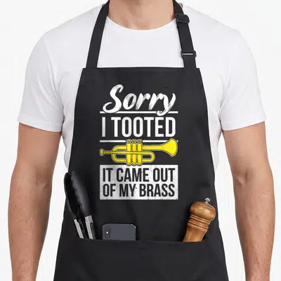 Player trumpet apron - Yellow cartoon trumpet with stacked white text reading Sorry I Tooted It Came Out Of My Brass — shown on a black cotton canvas apron against a white background