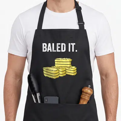 Pun farmer apron - Baled It text above four stacked yellow cartoon hay bales — shown on a black cotton canvas apron against a white background