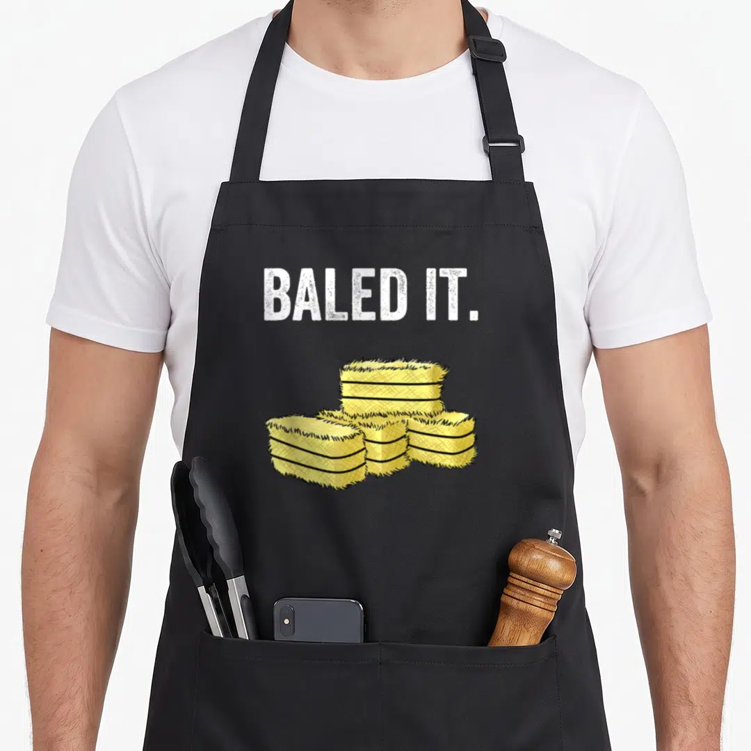 Pun farmer apron - Baled It text above four stacked yellow cartoon hay bales — shown on a black cotton canvas apron against a white background