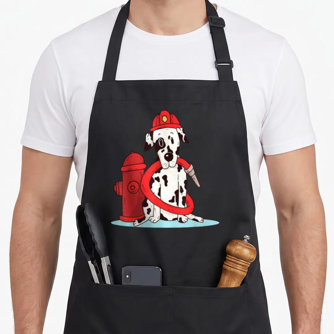 Firefighter dog apron - Cartoon dalmatian in red helmet holding hose beside red fire hydrant — shown on a black cotton canvas apron against a white background