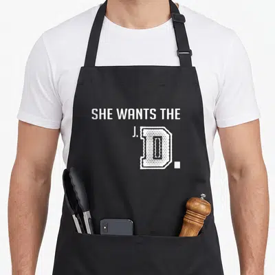 She student aprons - White text reading SHE WANTS THE J.D. with a large textured D on a grey background — shown on a black cotton canvas apron against a white background