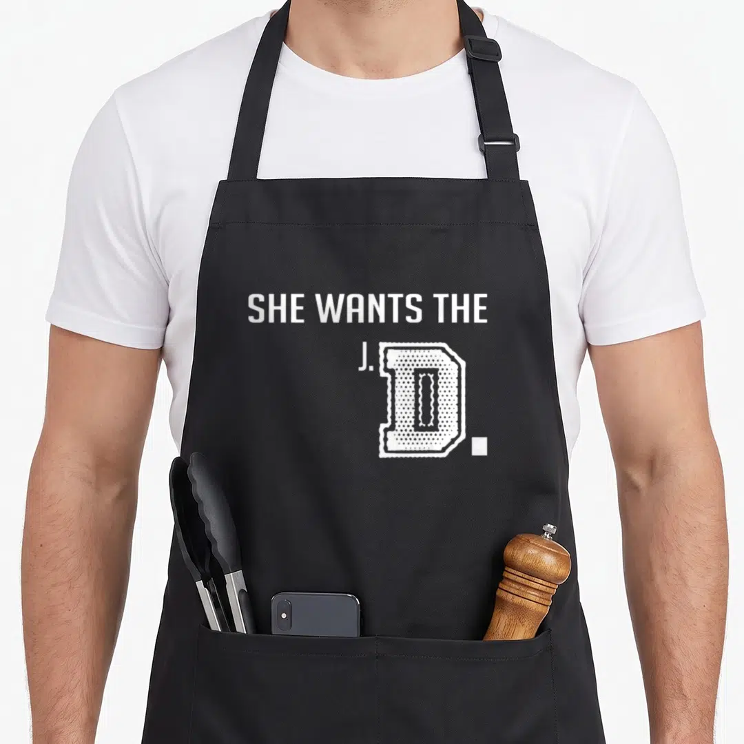 She student aprons - White text reading SHE WANTS THE J.D. with a large textured D on a grey background — shown on a black cotton canvas apron against a white background
