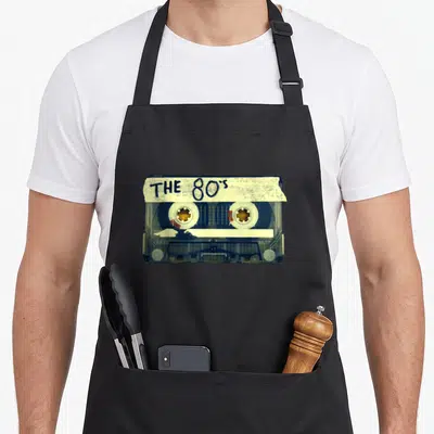 Mix 80s apron - Realistic cassette tape with THE 80's handwritten label in blue and off-white — shown on a black cotton canvas apron against a white background