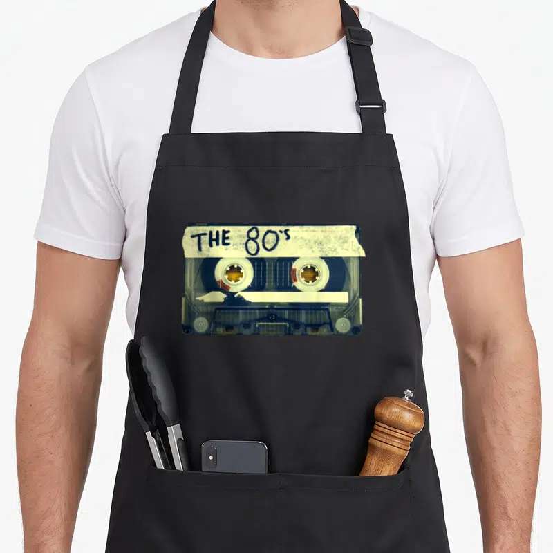 Mix 80s apron - Realistic cassette tape with THE 80's handwritten label in blue and off-white — shown on a black cotton canvas apron against a white background