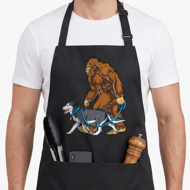 Bigfoot husky apron - Cartoon brown bigfoot walking a grey and white husky on a blue leash — shown on a black cotton canvas apron against a white background