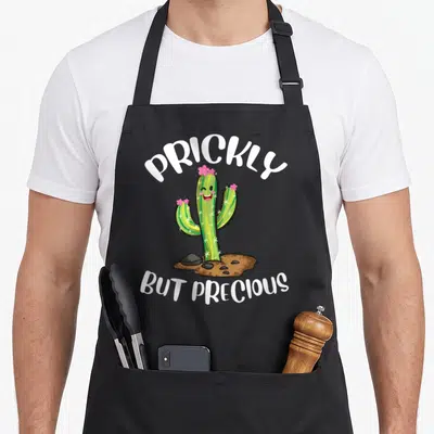 Prickly cactus apron - Smiling cartoon cactus with pink flowers and white PRICKLY BUT PRECIOUS text over brown dirt — shown on a black cotton canvas apron against a white background