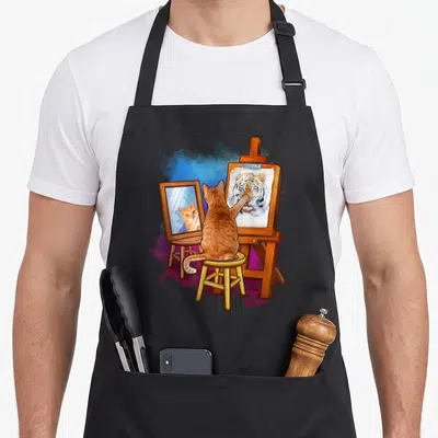 Cat artistic apron - Orange cat painting tiger portrait on easel with mirror reflection in cartoon style — shown on a black cotton canvas apron against a white background