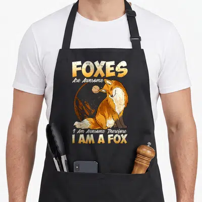 Cool fox print apron - Cartoon fox sitting before moon and mountains with orange yellow white text — shown on a black cotton canvas apron against a white background