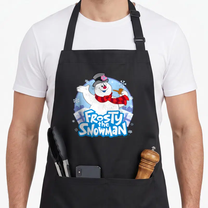 Funny frosty snowman apron - Waving cartoon snowman with red scarf and Frosty the Snowman text over two gift boxes — shown on a black cotton canvas apron against a white background