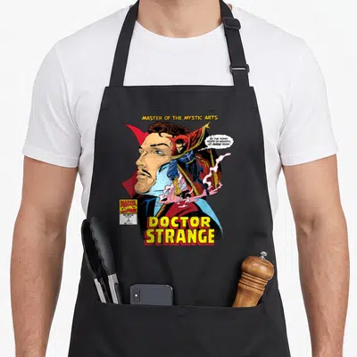 Comic doctor apron - Doctor Strange comic cover with large face, full body figure, and yellow title text — shown on a black cotton canvas apron against a white background