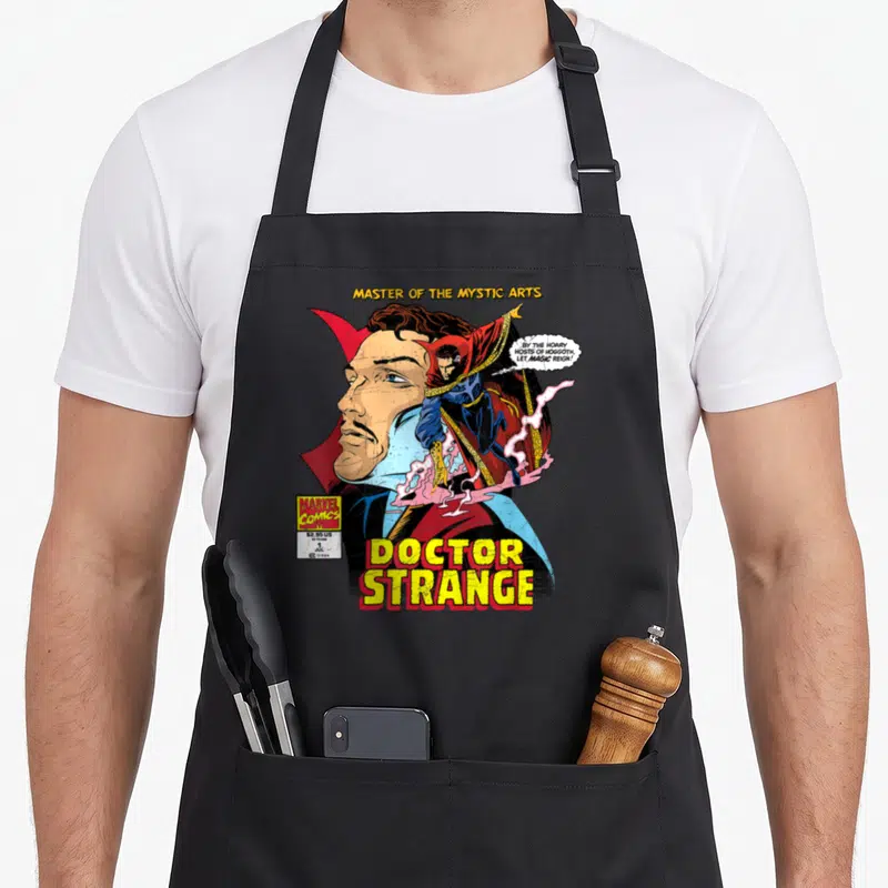 Comic doctor apron - Doctor Strange comic cover with large face, full body figure, and yellow title text — shown on a black cotton canvas apron against a white background