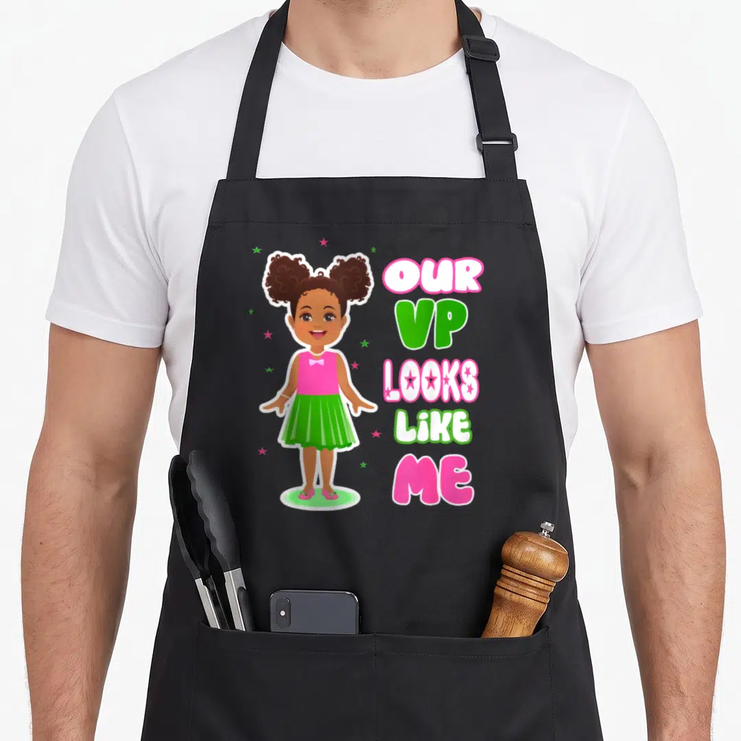 Our aka apron - Cartoon girl in pink top and green skirt beside stacked text reading OUR VP LOOKS LIKE ME with stars — shown on a black cotton canvas apron against a white background