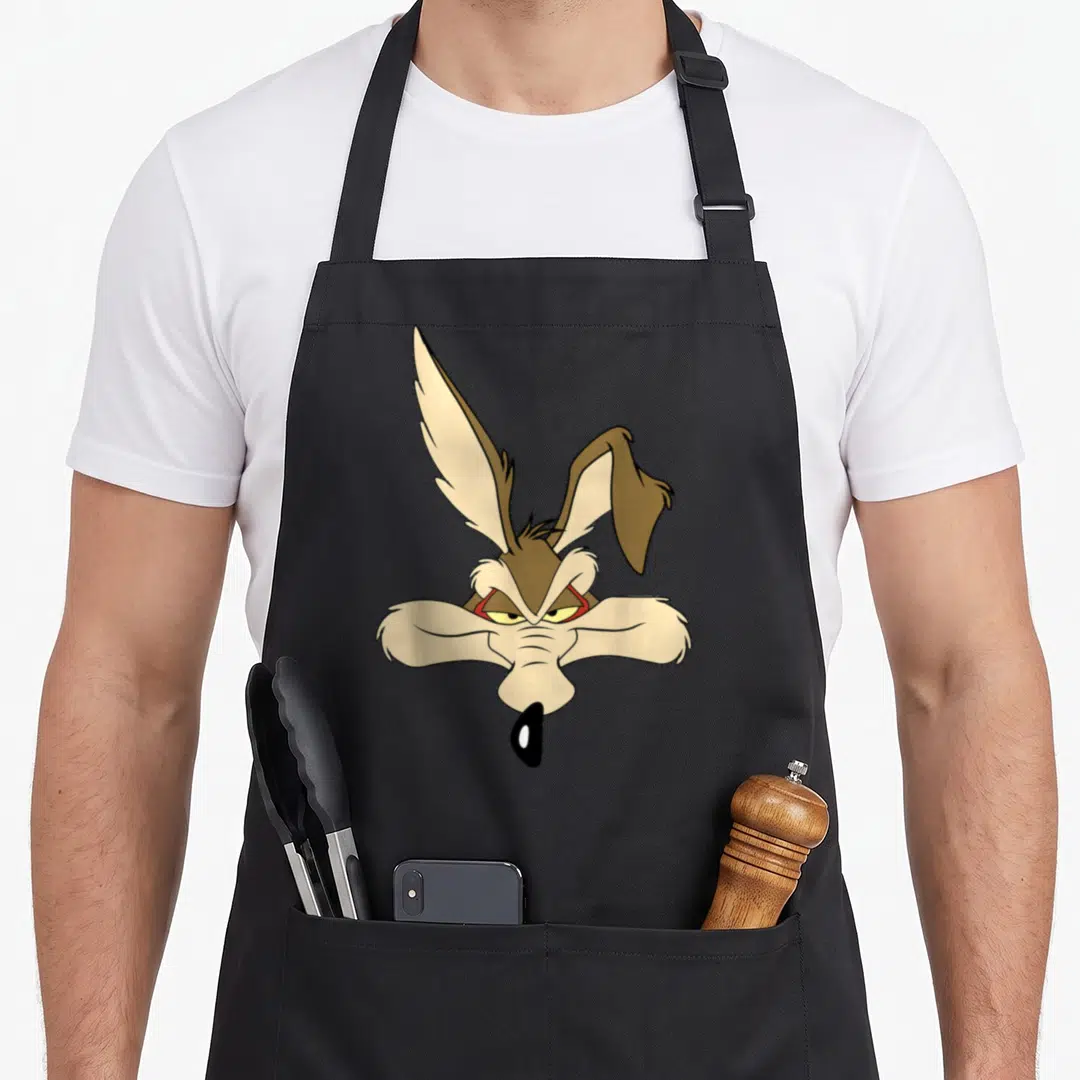 Cute wile e.a apron - Brown and tan cartoon coyote head illustration with one upright ear and one flopped ear — shown on a black cotton canvas apron against a white background