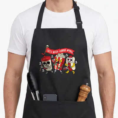 Watch skeleton apron - Three cartoon snacks holding weapons under a red banner reading LET'S WATCH HORROR MOVIES — shown on a black cotton canvas apron against a white background