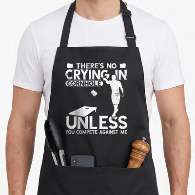 Crying game aprons - Distressed white silhouette of person throwing bean bag with cornhole board and sarcastic text — shown on a black cotton canvas apron against a white background